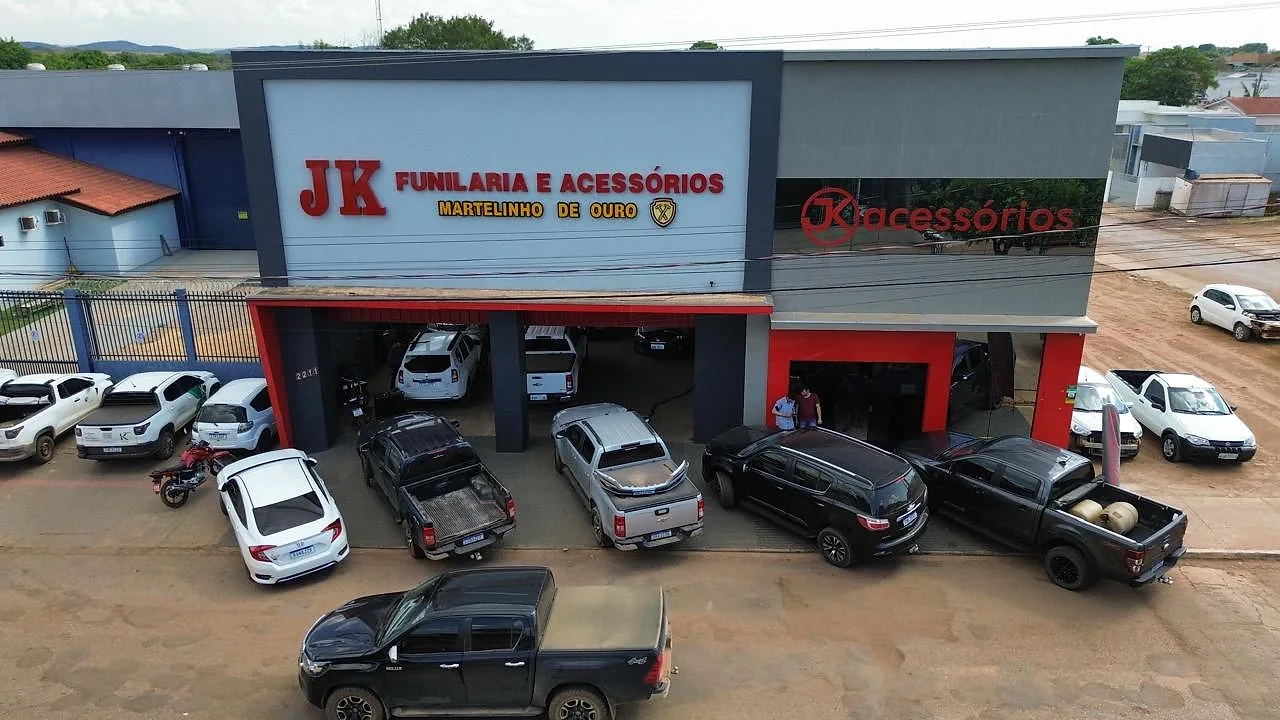 Vista aérea da fachada da JK Funilaria e Acessórios na Av. Brasil, 2211, em Paranatinga – MT: a oficina com a placa "Martelinho de ouro" e, ao lado, a loja JK Acessórios, com carros e picapes estacionados em frente.
